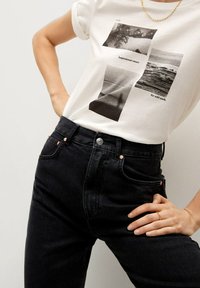 Biała koszulka z czarnym nadrukiem graficznym, zestawiona z wysokim stanem czarnymi dżinsami, które mają miedziane nity i gładką teksturę denimu.
