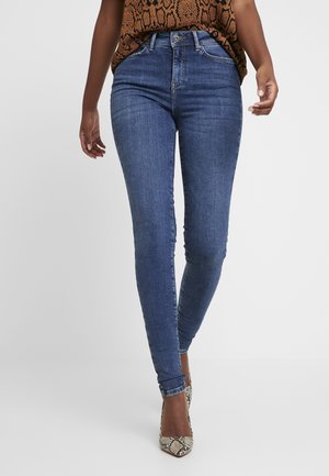 Kobieta ubrana w niebieskie rurki z wysokim stanem, brązowy top w wężowy wzór i szpiczaste szpilki z wężowym nadrukiem, stojąca na jednolitym tle.