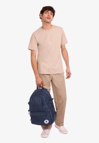 Blå ryggsäck med flera fack och blixtlås, tygtextur och en rund logotypmärke. Modell bär beige skjorta och ljusa byxor.