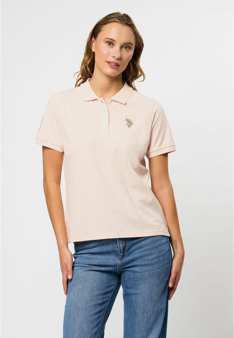 Lichtroze polo shirt met een klassieke kraag, drie knopen en een gouden geborduurd logo op de borst. Korte mouwen en van katoen.