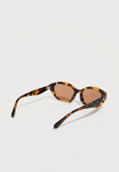Lunettes de soleil à motif écaille de tortue avec des verres ovales marron et des branches fines, posées ouvertes sur une surface blanche.