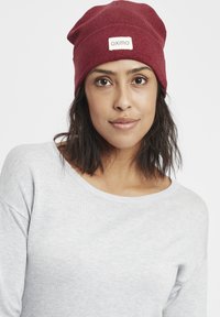 Maroon knit beanie with a folded cuff and a white label. Worn with a light gray sweater featuring a wide neckline. Soft texture.