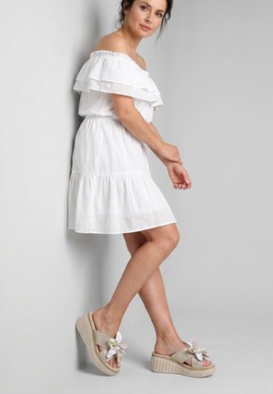 Frau in weißem schulterfreiem Kleid, die an eine Wand lehnt, trägt beige Plateau-Sandalen mit Schleifendetails.