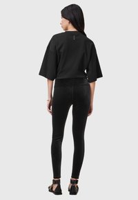 Donna con lunghi capelli scuri che indossa una blusa nera larga, leggings in velluto neri e sandali neri con fibbie dorate, in piedi di fronte a uno sfondo semplice.