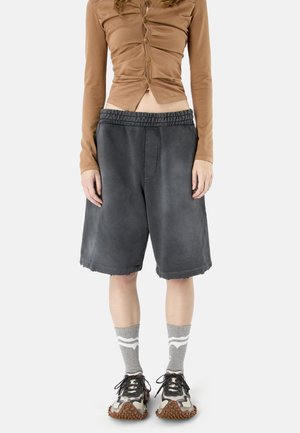 Persoon met een bruin kort topje met knopen, losse donkergrijze knielange korte broek, grijze sokken met witte strepen en meerkleurige sneakers.