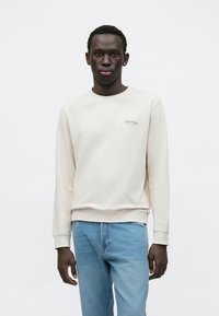 Φωτεινή μπεζ φούτερ με στρογγυλή λαιμόκοψη και ριμπ μανσέτες, με ραμμένη λεπτομέρεια λογότυπου στο αριστερό στήθος, σε συνδυασμό με ανοιχτό μπλε τζιν.