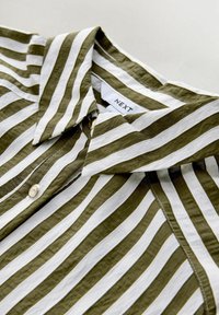 Proužkovaná košile s střídajícími se olivově zelenými a bílými pruhy, s límečkem na knoflíky a viditelnými stehmi. Štítek nese nápis "NEXT."