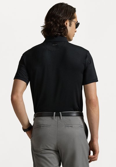 Svart kortärmad polo t-shirt med en slät yta, med en diskret logotyp på ryggen vid nacken. Tillsammans med grå byxor och ett svart bälte.