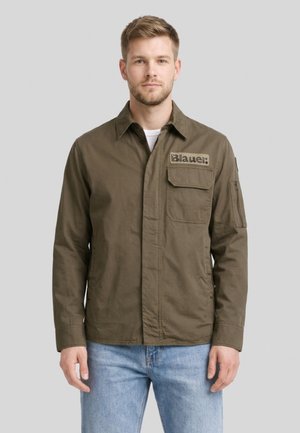 Mężczyzna w oliwkowo-zielonej zapinanej na guziki kurtce z klapą na piersi i naszywką „Blauer”, w połączeniu z jasnoniebieskimi dżinsami na jednolitym tle.