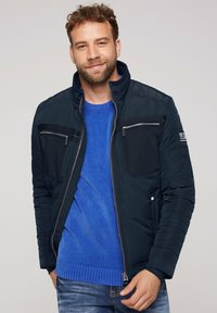 Mørkeblå zipped jakke med høj krave, polstrede skulderdetaljer og fremadskuende lynlås lommer. Båret over en blå ribbestrikket sweater og jeans.