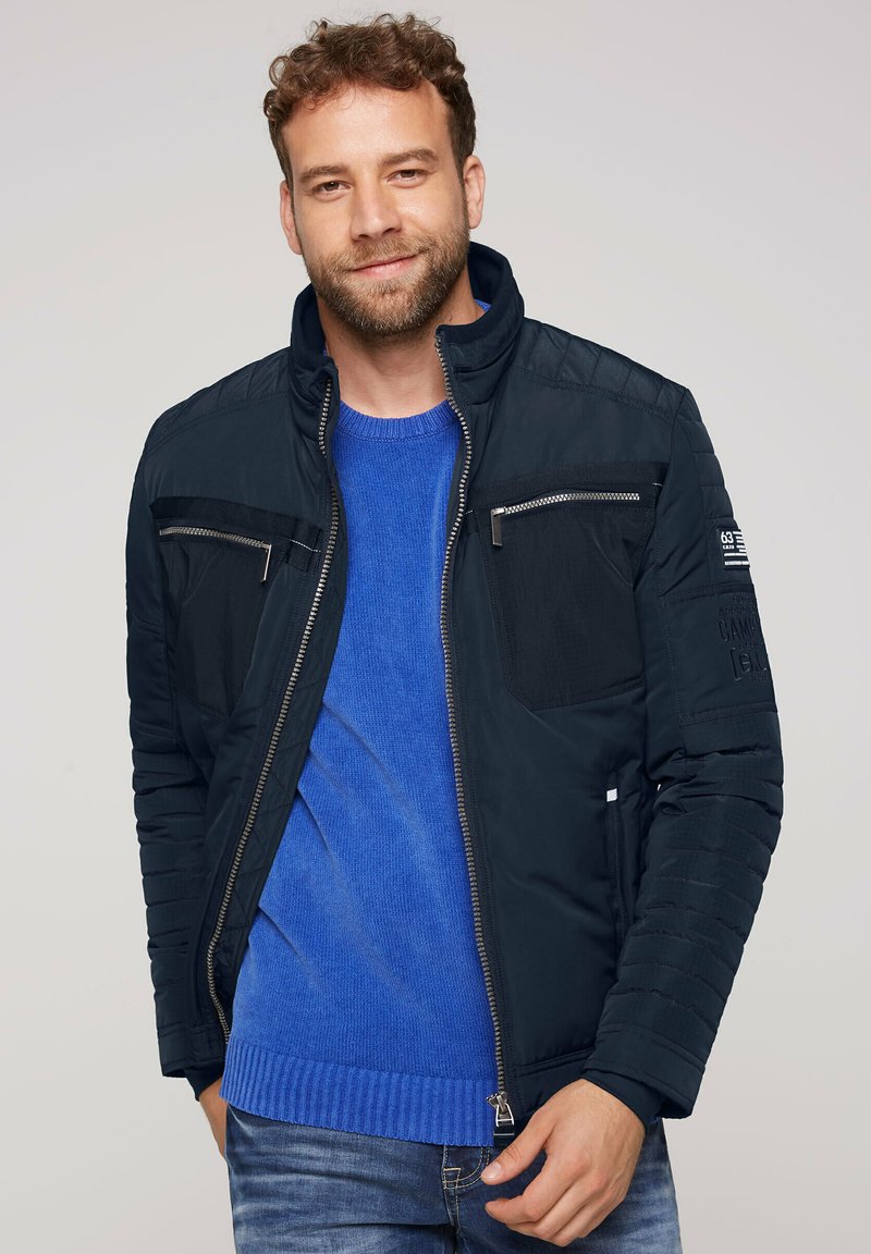 Mørkeblå zipped jakke med høj krave, polstrede skulderdetaljer og fremadskuende lynlås lommer. Båret over en blå ribbestrikket sweater og jeans.