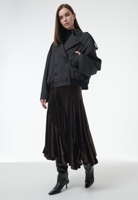 Chaqueta corta negra de doble botonadura con mangas voluminosas y cinturilla ajustada, combinada con una falda maxi de terciopelo marrón en capas y botas negras hasta la rodilla.