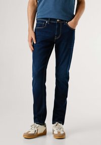 Tmavomodré rifle so slim fit strihom, s kontrastným šitím, piatimi vreckami a lesklým kovovým gombíkom. Skombinované s bielymi a pieskovými teniskami.