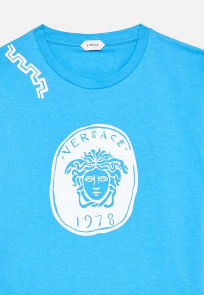 Μπλε t-shirt Versace με το λογότυπο κεφαλής Μέδουσας και το κείμενο "VERSACE 1978" σε λευκό στο εμπρόσθιο μέρος, και λευκό γεωμετρικό σχέδιο κοντά στον γιακά.