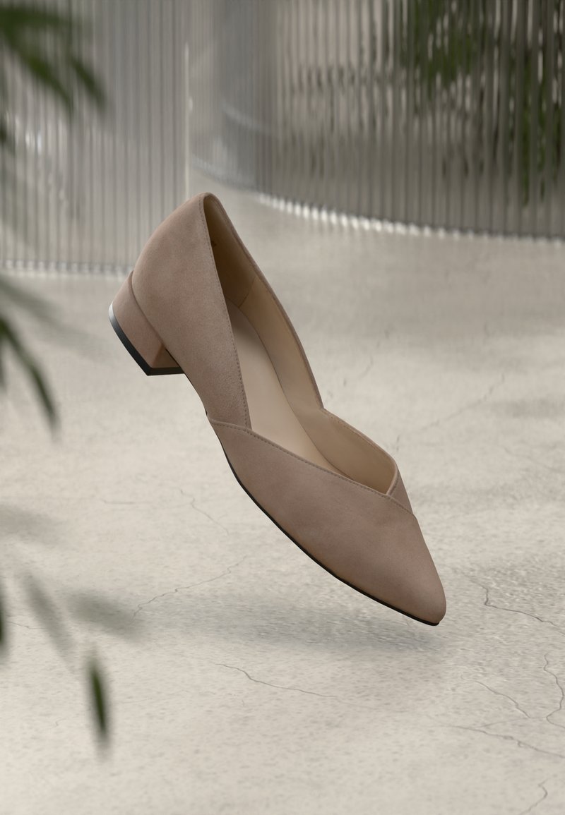Chaussure basse pour femme en suède beige à bout pointu flottant au-dessus d'un sol clair fissuré avec en arrière-plan des plantes floues et un mur texturé.