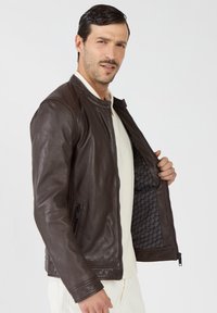 Bruine leren jas met ritssluiting en geribbelde schouders. De binnenkant heeft een gemêleerde voering. Model poseert met de jas open.