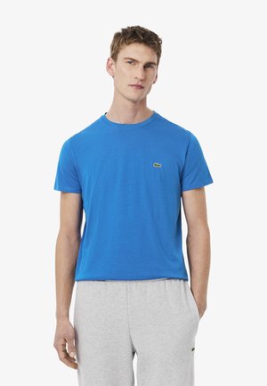 Korte mouwen blauwe katoenen T-shirt met een ronde hals en een klein groen logo accent op de linker borst. Gecombineerd met grijze sweatpants.