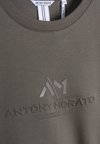 Camiseta slim fit verde oliva con logo elevado de caucho "ANTONY MORATO" y "ESTABLECIDO DOS MIL SIETE" en negro. Textura suave.