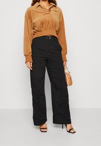 Braunes Corduroy-Cropped-Sweatshirt, schwarze Weitbein-Hosen mit Raffung und schwarze Sandalen. Begleitet von einer kleinen beigen Handtasche.
