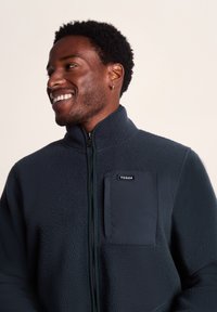 Dark blue fleece jacket with a high collar, zip front, and a contrasting pocket. Soft texture with stitching details and logo on the chest.