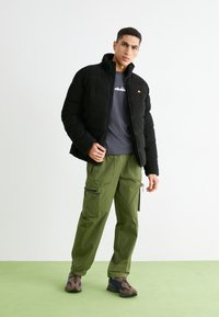 Schwarze Pufferjacke mit gestepptem Muster, hohem Kragen und orangefarbener Logo. Kombiniert mit olivgrünen Cargo-Hosen mit mehreren Reißverschlüssen.