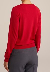 Röd stickad tröja med långa ärmar och rund halsringning, parat med grå byxor. Tyget verkar mjukt med en slät textur.