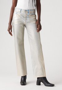 Jeans larghi in denim metallico chiaro con una texture lucida, caratterizzati da cuciture azzurre e design a cinque tasche, abbinati a stivali neri alla caviglia.