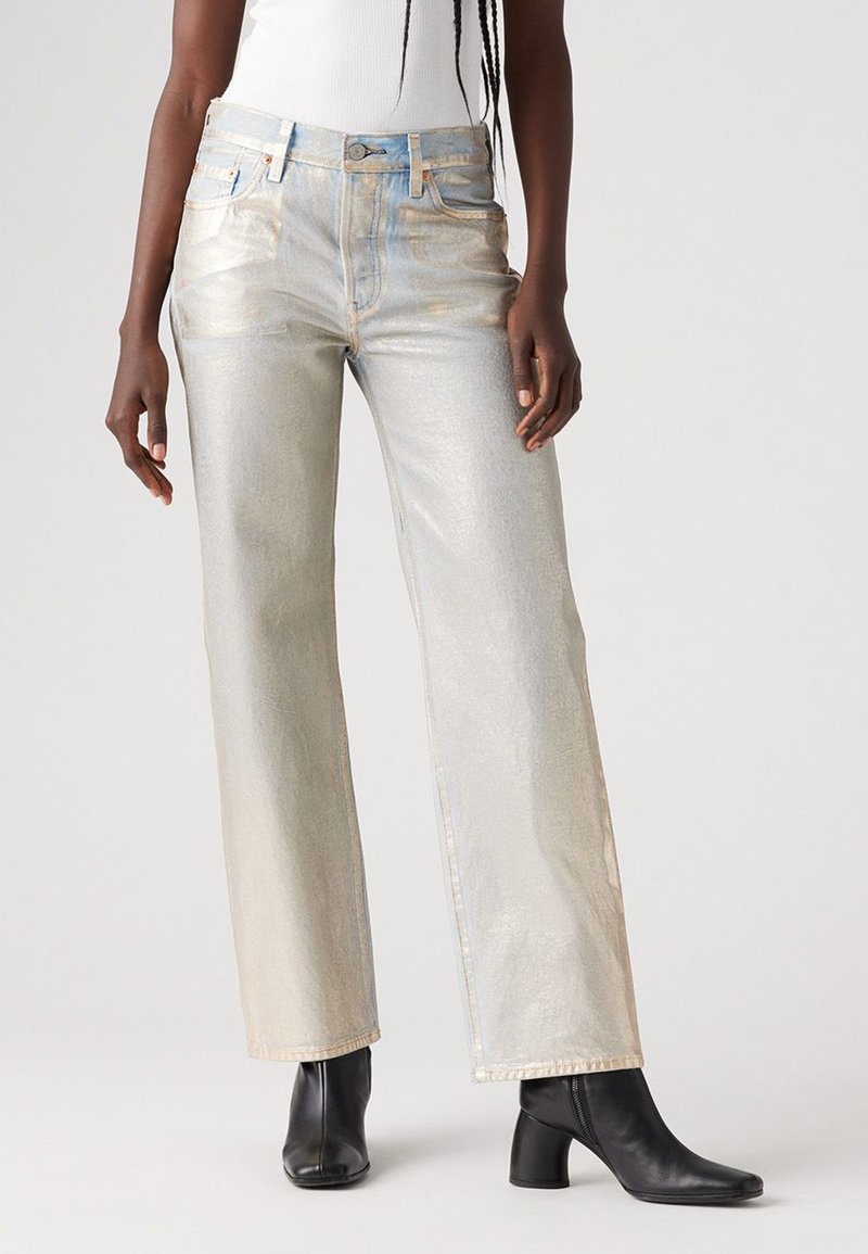 Jean large en métal léger avec une texture scintillante, ornés de coutures bleu clair et d'un design à cinq poches, associés à des bottines noires.