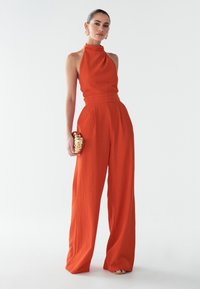 Oranžový overal s halterneck výstrihom, priliehavým telom a širokými nohavicami, vyrobený z texturovanej tkaniny. Doplněný zlatou kabelkou.