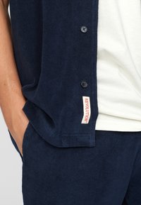 Marinblå frottéknappad skjorta med avslappnad passform, med en ficka och en röd "REVOLUTION"-etikett på sidan. Snygg textur.