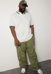 Kortärmad vit polo med flerfärgad kant, olivgröna cargobyxor och vita sportskor. Bär en olivgrön baseballkeps.