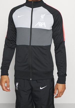 Mężczyzna ubrany w czarno-szarą kurtkę na zamek Liverpool FC oraz czarne spodnie Nike z widocznymi logo Liverpool i Nike.