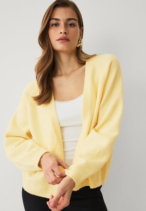 Kvinde justerer ærmet på gul cardigan over hvid top, iført guldbøjle, mod ensartet lyst baggrund.