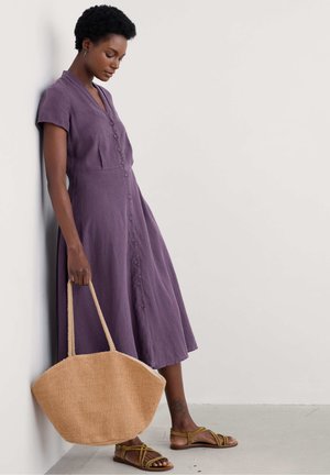 Lila Hemdkleid mit kurzen Ärmeln, fließendem Rock, strukturiertem Stoff; kombiniert mit einer großen gewebten beigen Tasche und braunen Riemensandalen.