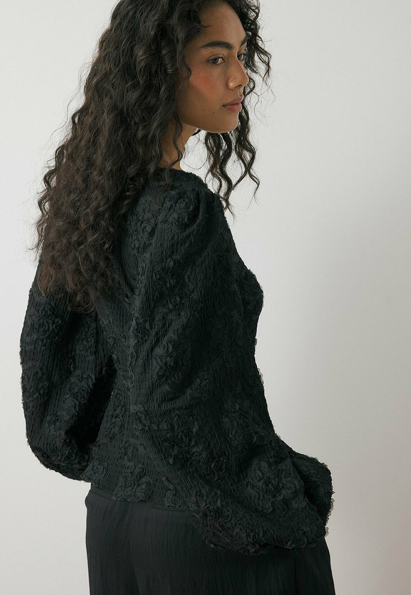 Zwarte textuur top met bloemenpatroon, puffed sleeves en een getailleerde pasvorm. Beschikt over een zachte, gekreukte stof.