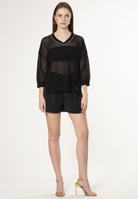 Maglione nero a maglia con un design a rete aperta e morbida, scollo a V e maniche a tre quarti, abbinato a pantaloni shorts neri e sandali con tacco dorati.
