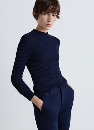 Giovane con capelli corti che indossa un maglione a coste scuro e pantaloni scuri abbinati con le mani in tasca contro uno sfondo semplice.