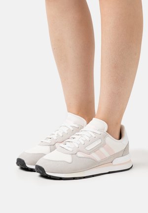 Baskets blanches et gris clair avec des rayures roses portées par une personne debout sur une surface blanche.