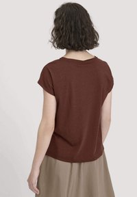 Frau mit kurzen lockigen Haaren, die ein braunes kurzärmliges Oberteil und einen beigen Rock trägt, von hinten an einer einfarbigen hellen Hintergrund.