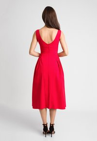 Czerwona sukienka bez rękawów z niskim dekoltem na plecach, plisowana spódnica i dopasowany gorset, wykonana z gładkiego materiału, zestawiona z czarnymi szpilkami.