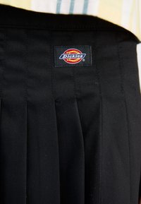 Melnas plisētās bikses, izgatavotas no izturīga auduma ar redzamu "Dickies" logotipa birku. Īpašības ietver gludu tekstūru un pielāgotu dizainu.