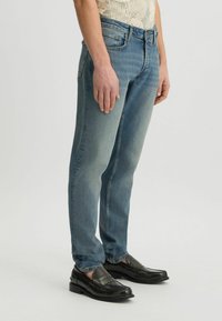 Jeans rectos desgastados de color azul, hechos de denim. Cuentan con un diseño clásico de cinco bolsillos y una cintura de altura media. Combinados con mocasines negros.