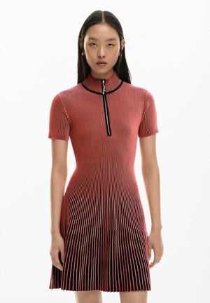 Žena v priliehavých červených krátkych rukávoch so zvislými čiernymi pruhmi a s polozipsovým výstrihom, stojaca na bielom pozadí.