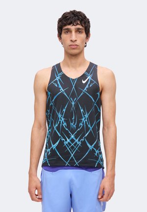 Junger Mann trägt ein schwarzes ärmelloses Nike-Oberteil mit blauen abstrakten Linien und hellblauen Sportshorts, steht vor einem einfarbigen Hintergrund.