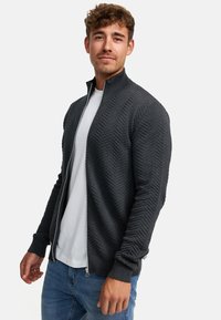 Un cárdigan con cremallera de color gris oscuro con un patrón de chevron texturizado, puños y dobladillo acanalados, usado sobre una camiseta blanca y combinado con jeans azules.