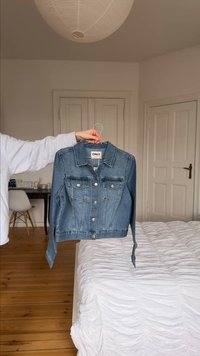 Kéz, amely kék farmerdzsekit tart fogason egy fehér ágy fölött egy minimál stílusú hálószobában, fa padlóval és zárt ajtókkal.