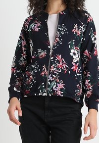 Vrouw in een marineblauwe zip-up jas met een roze, witte en groene bloemenpatroon, gecombineerd met zwarte broeken en een lichtroze shirt eronder.