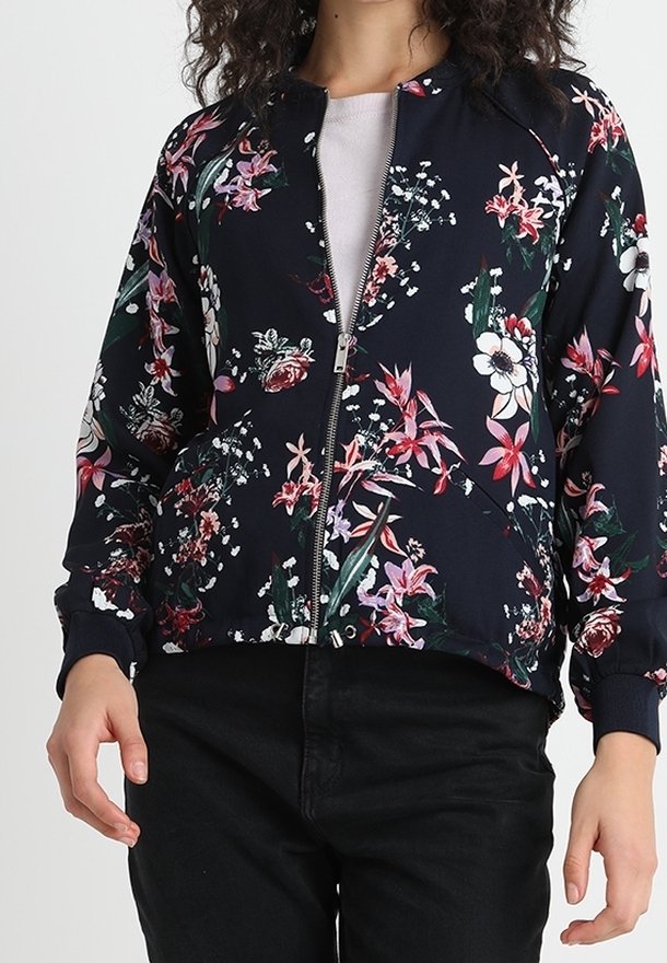Vrouw in een marineblauwe zip-up jas met een roze, witte en groene bloemenpatroon, gecombineerd met zwarte broeken en een lichtroze shirt eronder.