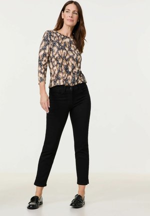 Woman standing with hands relaxed, wearing a brown and black patterned long-sleeve top, black slim pants, and black loafers on a light background.