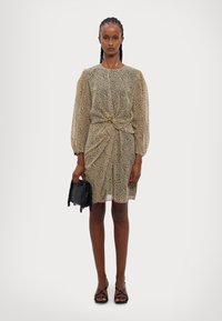 Krátke rukávy béžových šiat s čiernym leopardím vzorom, s naťahovacím detailom v oblasti pása, v kombinácii s malou čiernou kabelkou a čiernymi sandálmi na pásiky.